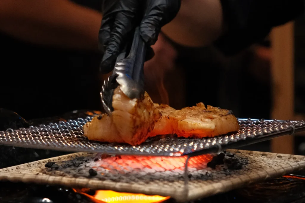 A person in black gloves using tongs to handle grilled food on a wire mesh over a flame.