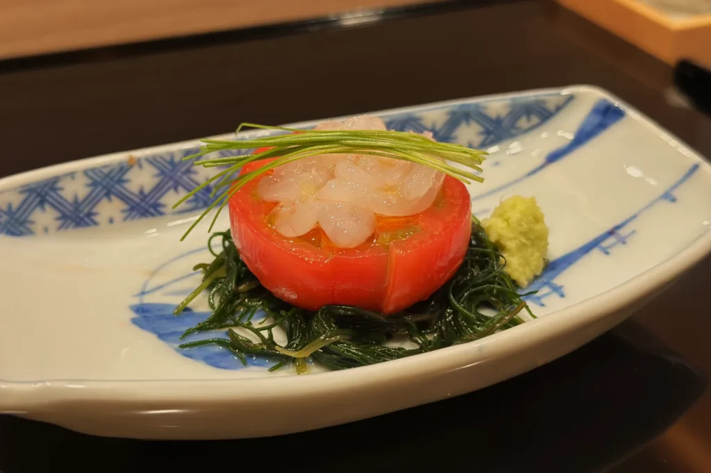 A plate of sliced tomato with translucent seafood on top, garnished with green onion and served on a bed of seaweed, accompanied by a dollop of wasabi.