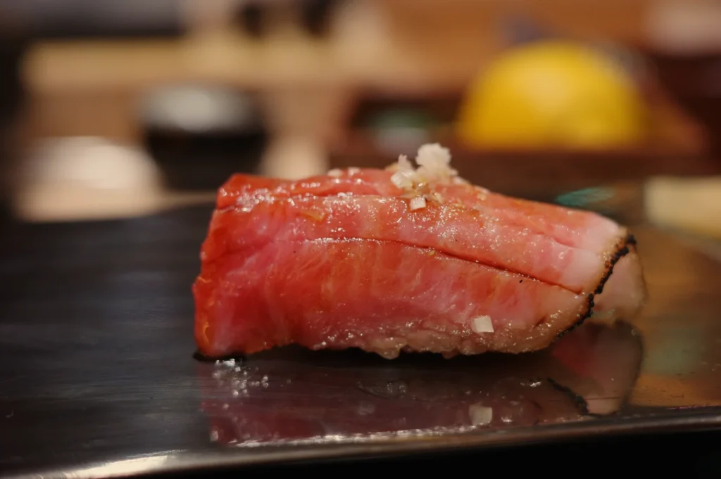 A glossy slice of seared tuna nigiri topped with a small garnish rests on a dark plate, with a softly blurred sushi counter creating an intimate dining mood.