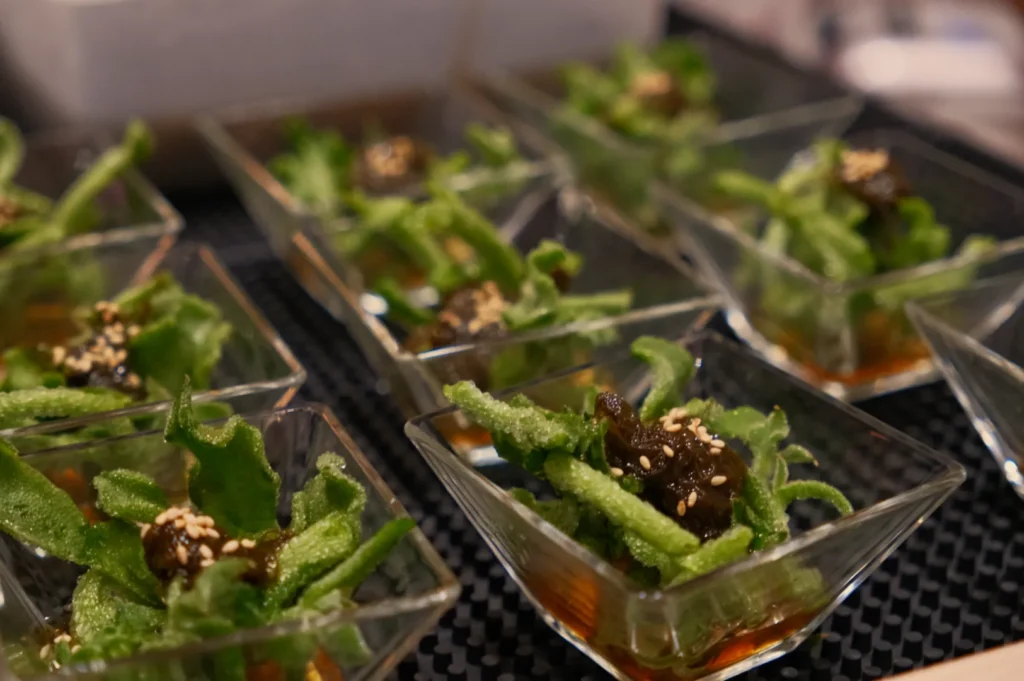 Small dishes containing green vegetables topped with sesame seeds, arranged on a black tray.