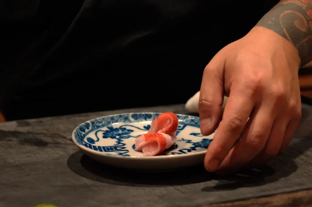 Chef Masa's hand placing a piece of sushi on a small blue and white plate.