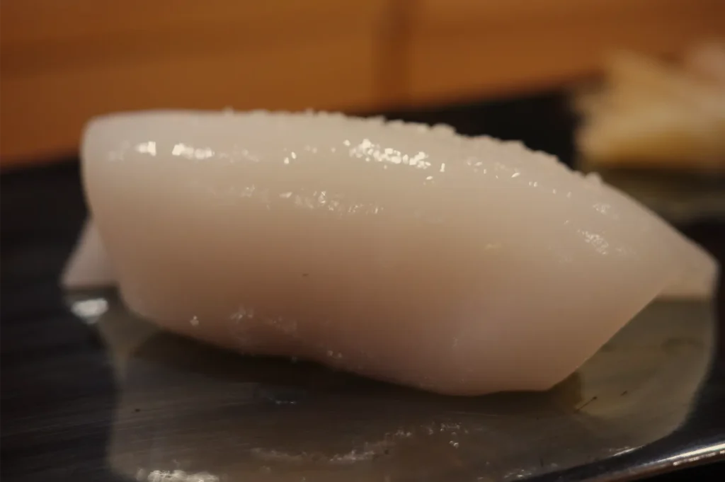 Glossy white squid nigiri sushi rests on a dark plate, its smooth, translucent texture highlighted against a warm, softly blurred background.