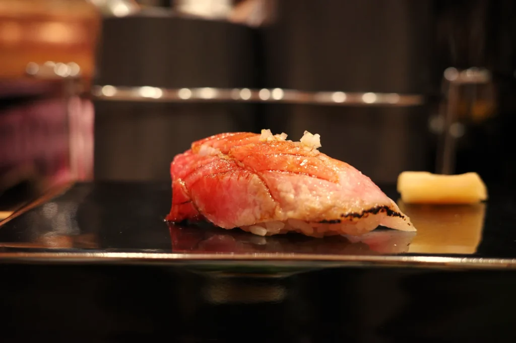 A piece of sushi, featuring a slice of fish on top of a small mound of rice, with a thin slice of ginger on the side.