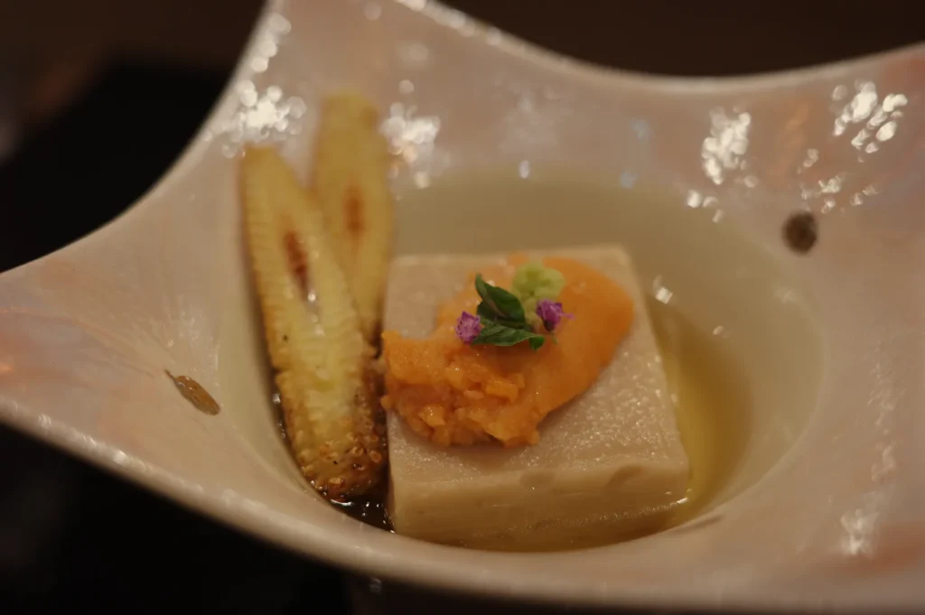 Gourmet appetizer on a spoon-shaped dish: a creamy tofu cube topped with orange purée and herbs, beside a grilled banana slice.