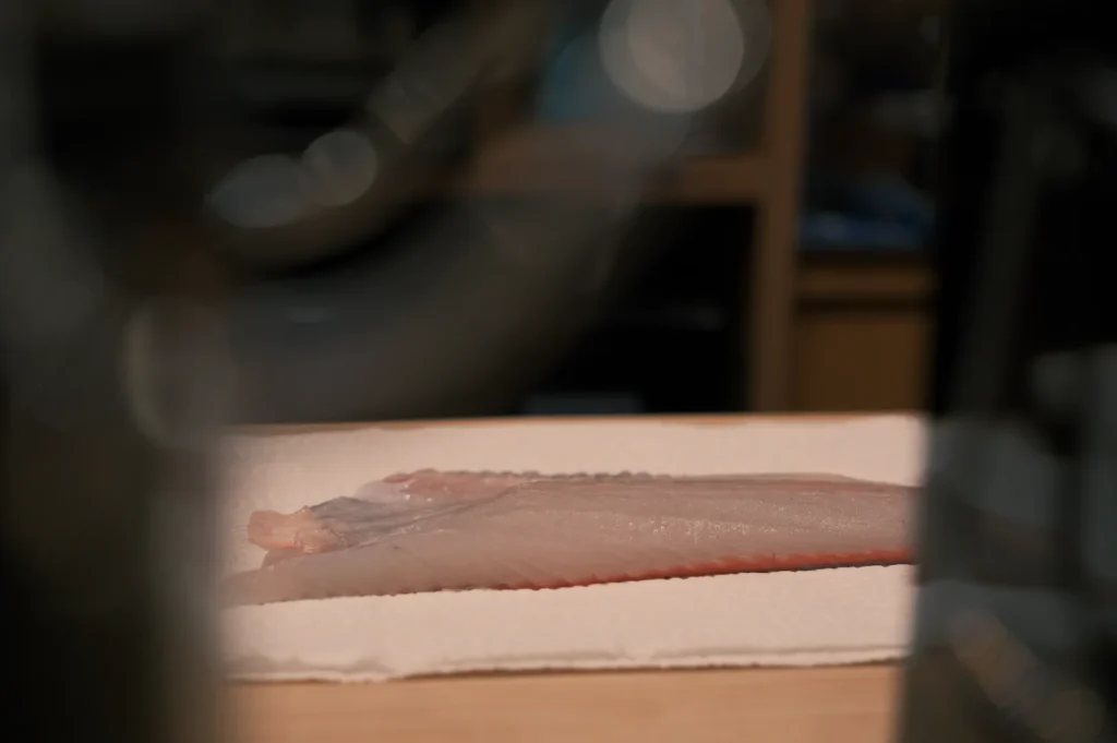 Raw fish fillet rests on a white cutting board in a dim kitchen, with blurred cookware in the background creating a quiet, subdued mood.