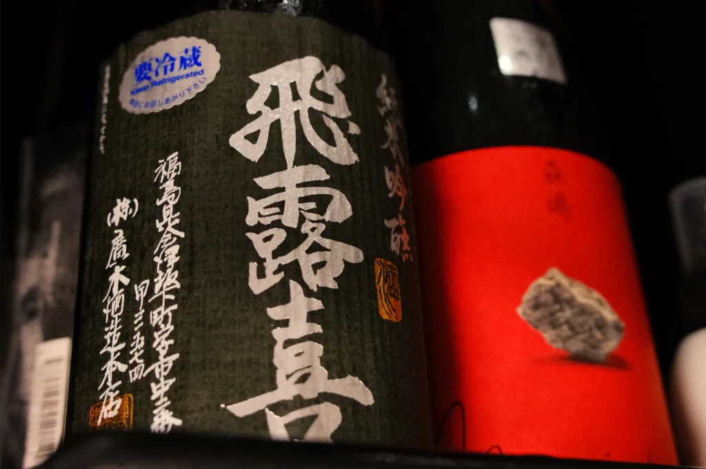 Close-up of dark sake bottles with bold Japanese calligraphy labels, a bright red label, and a textured stone-like emblem; moody lighting.