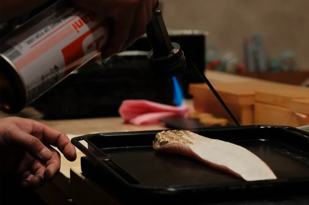 Chef Masa using a blowtorch to sear a piece of fish on a tray.