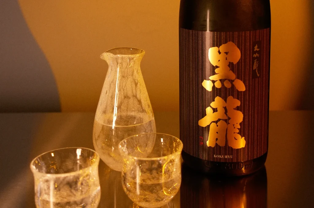 A bottle of sake labeled "黒龍" (Kokuryu) beside a clear glass sake carafe and two small glasses on a reflective surface.