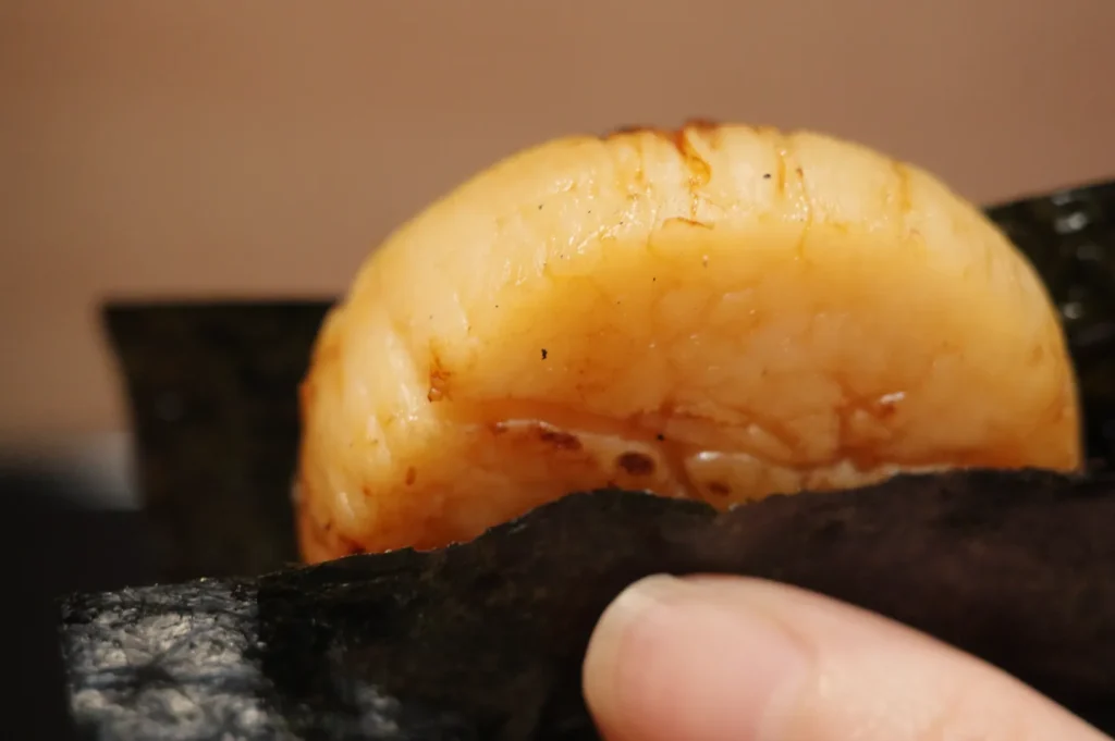 A close-up image of a piece of sushi, featuring a shiny, cooked seafood topping on a small bed of rice, held by fingers with a dark seaweed wrap.
