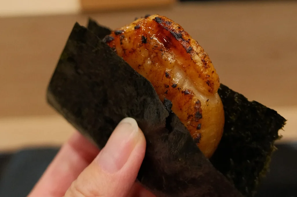 A piece of sushi wrapped in nori, featuring grilled seafood, possibly scallops, held in a person's hand.