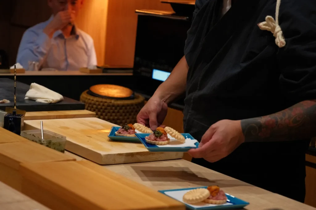 A chef holding plates of sushi in a restaurant, with a diner seated in the background.