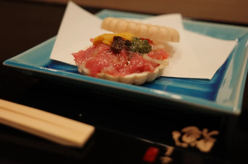 Close-up of a Japanese seafood appetizer on a blue plate, topped with minced red fish, yellow and green garnishes, and a crisp shell.