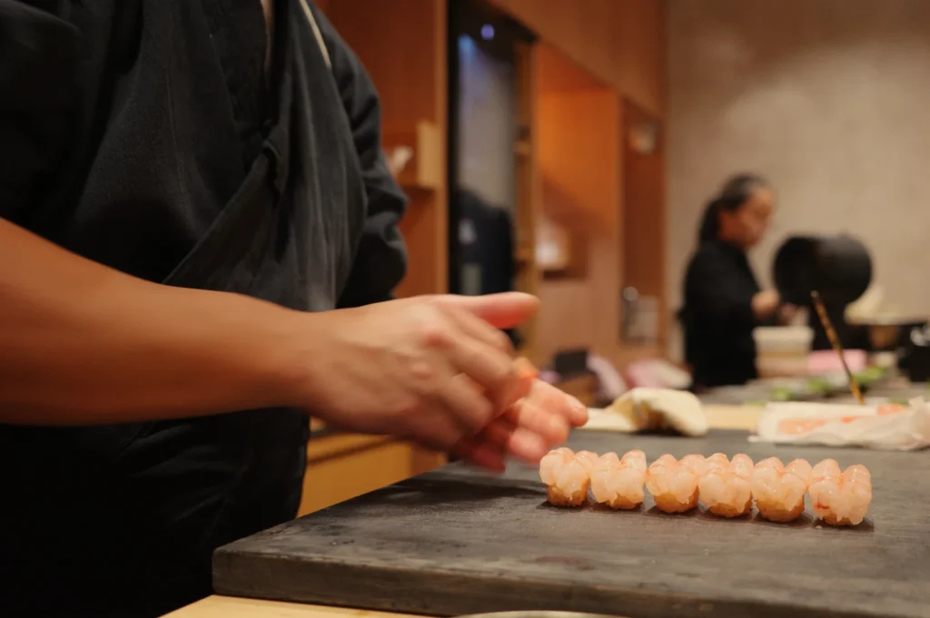 A sushi chef in a dark uniform arranges a neat row of salmon-topped sushi on a board, while another chef works in the warm, busy kitchen behind.