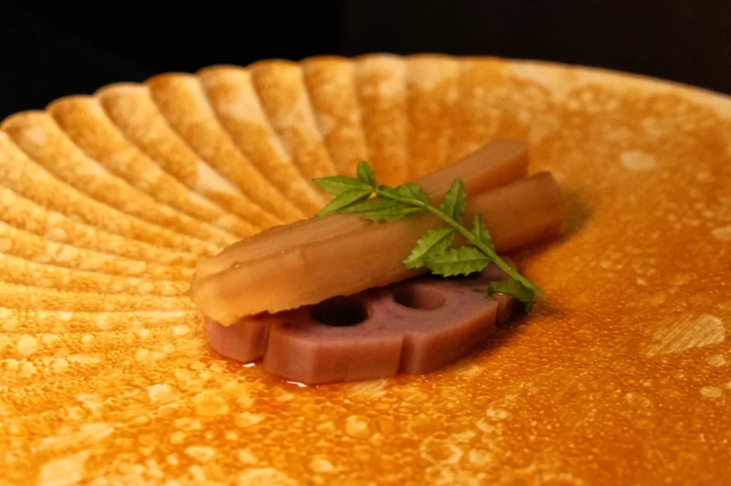 A plate with a decorative design featuring two pieces of food, one resembling a light-colored stick and the other a purple lotus root slice, garnished with a green herb.