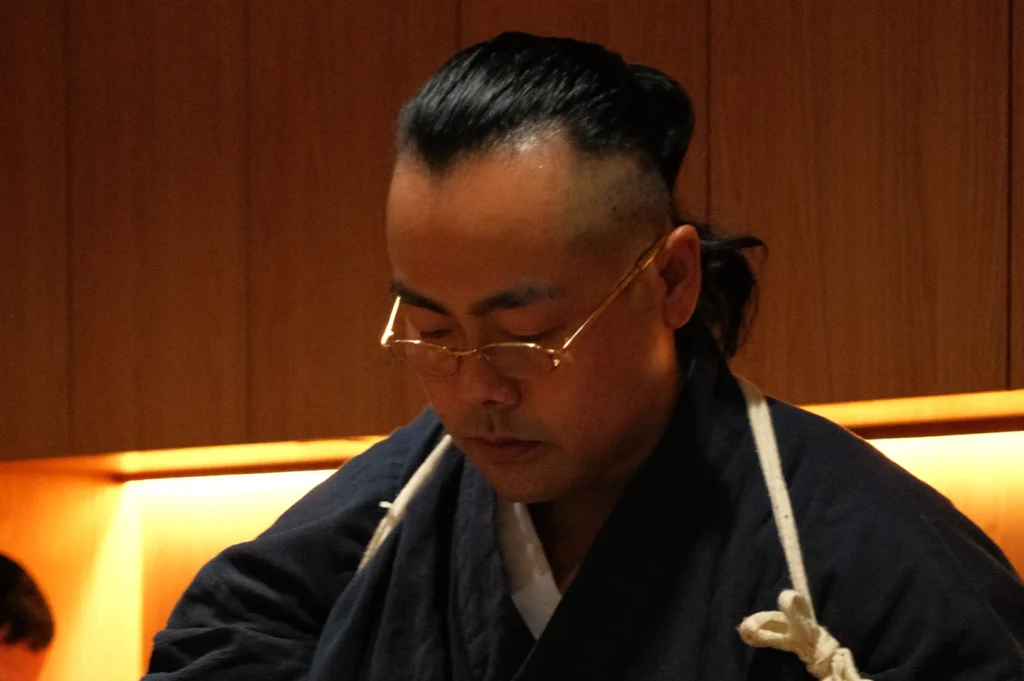A man wearing a navy blue traditional outfit with glasses, looking down while concentrating in a warmly lit wooden environment.