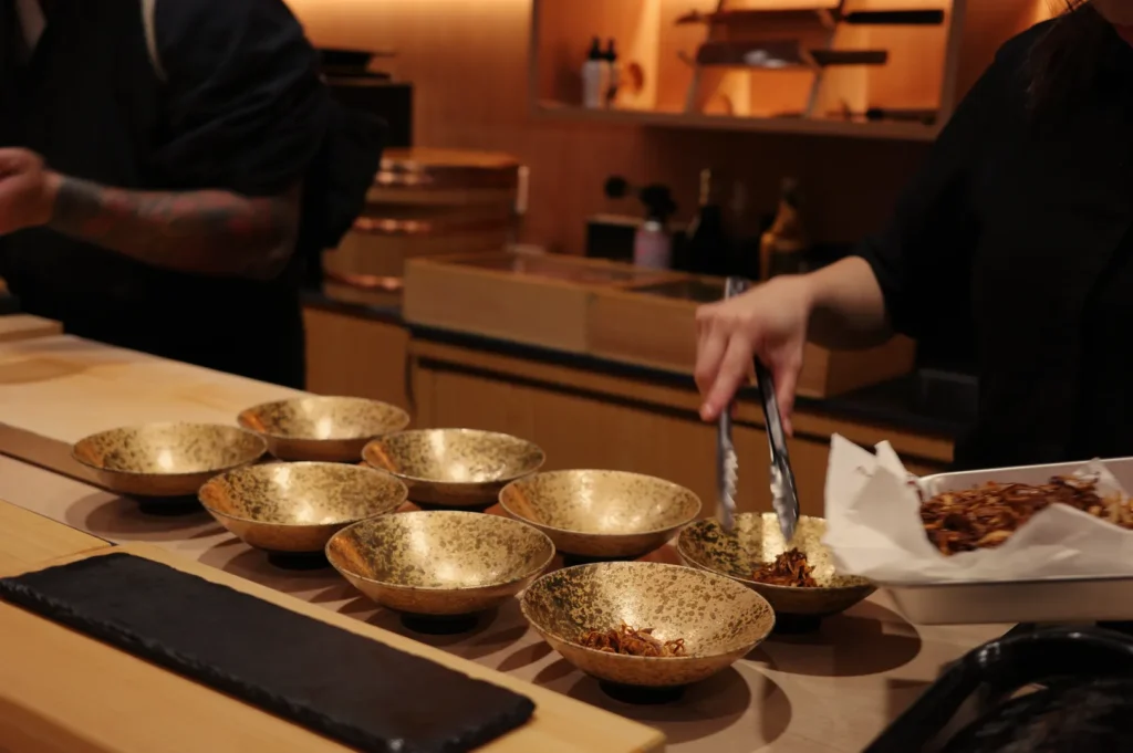 A person placing food into gold bowls on a wooden counter, with additional bowls and kitchen items in the background.