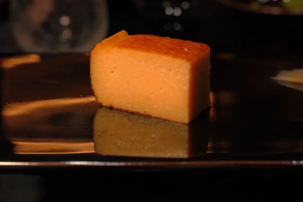 A rectangular piece of yellowish-orange cake on a dark plate, with a glossy surface reflecting the cake's shape.