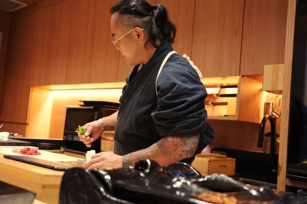 A chef preparing sushi in a modern kitchen setting, wearing a dark blue chef's uniform and showcasing tattoos on his arms.