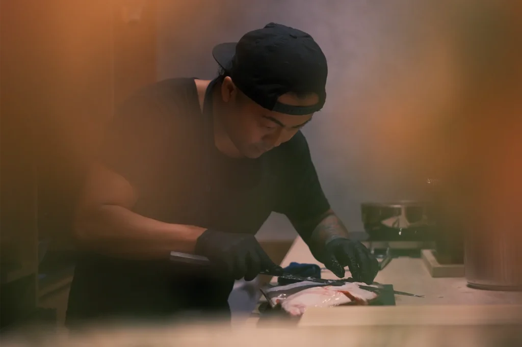 A focused chef in black clothing and gloves slices fish at a kitchen counter, surrounded by warm, hazy light in an intimate atmosphere.