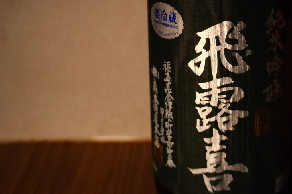Close-up of a black Japanese sake bottle with bold white kanji, red and white side lettering, and a blue seal on a warm wooden table.