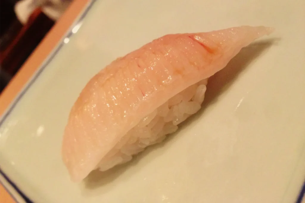 A close-up of pale pink nigiri sushi, with a glossy slice of fish draped over compact white rice on a cream-colored plate; simple, appetizing presentation.