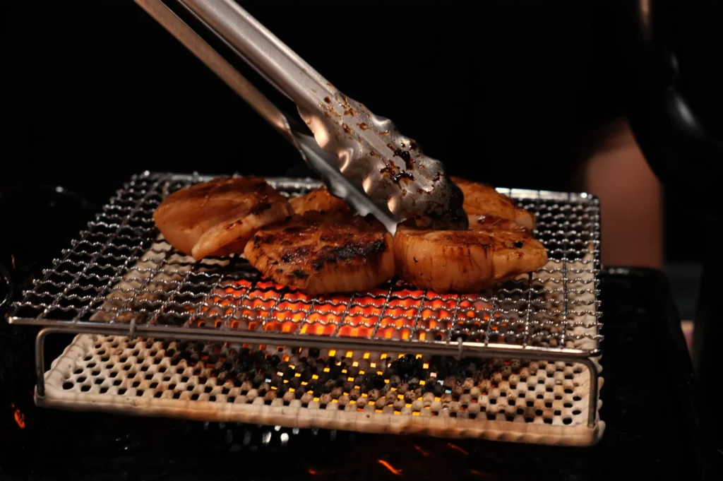 Grilled seafood pieces being flipped with tongs on a wire rack over glowing embers.