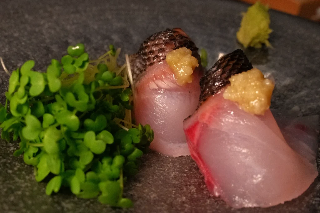 Two pieces of sushi with slices of fish topped with a dollop of condiment, placed on a black plate alongside fresh greens.