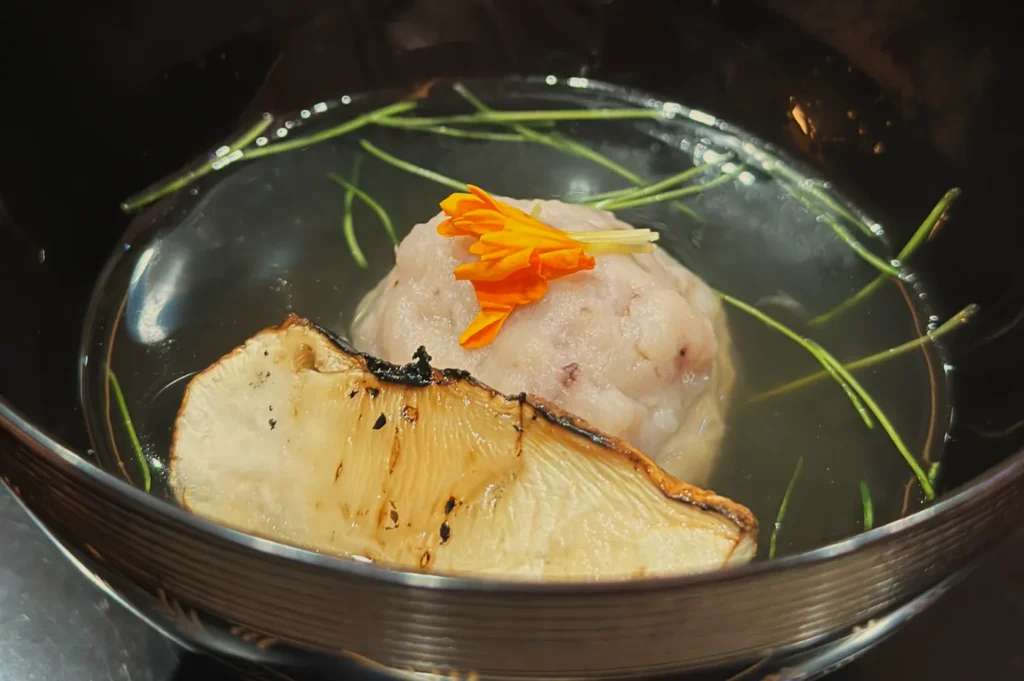 A bowl of clear broth with a round dumpling, garnished with a flower and herbs, alongside a slice of grilled mushroom.