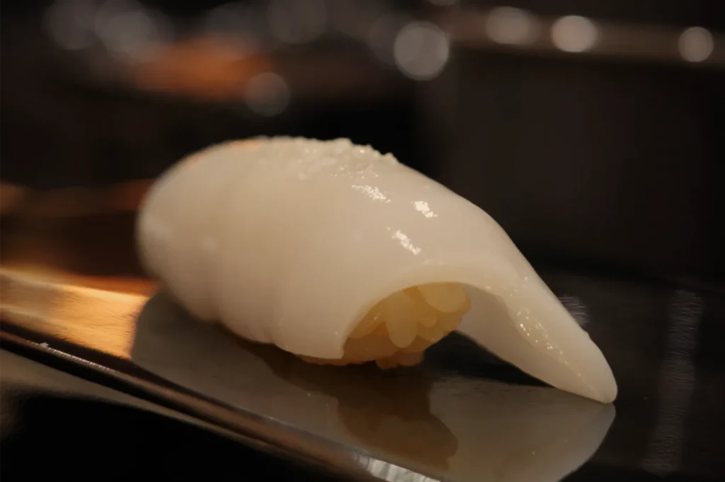 Close-up of glossy white squid nigiri sushi on a dark plate, its translucent topping draped over rice in warm, intimate lighting.