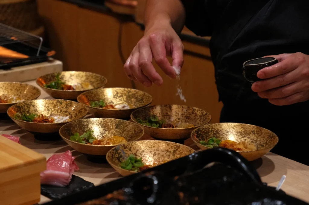 Chef Masa sprinkling seasoning over multiple gold bowl dishes arranged on a table.
