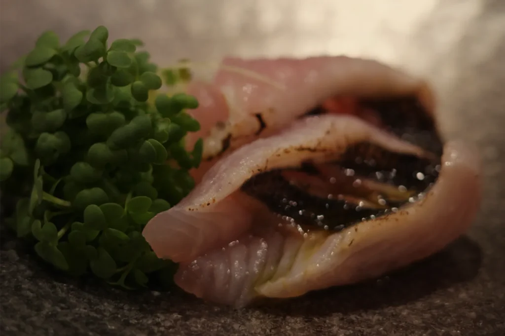 Close-up of an open raw shellfish with pink, glossy flesh and a dark, briny interior, served beside fresh green microgreens on a dark surface.