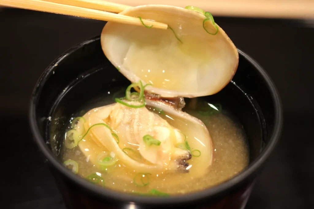 A bowl of miso soup with clams and chopped green onions, with a clam shell being lifted by chopsticks.