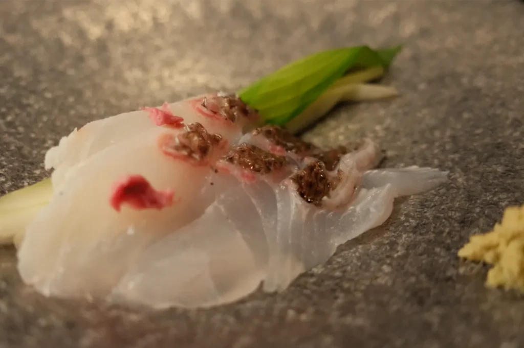 Close-up of translucent white sashimi topped with scallions, red garnish, and brown seasoning on a dark plate, with ginger nearby.