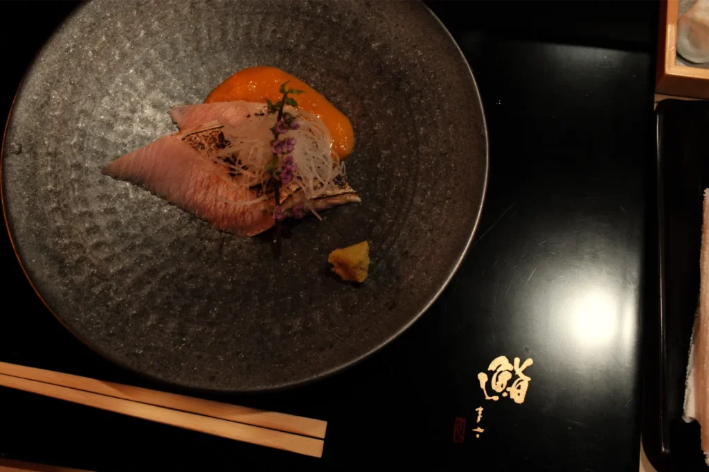A plate with sushi featuring fish slices, garnished with herbs and served with a sauce, placed on a textured black surface with chopsticks beside it.