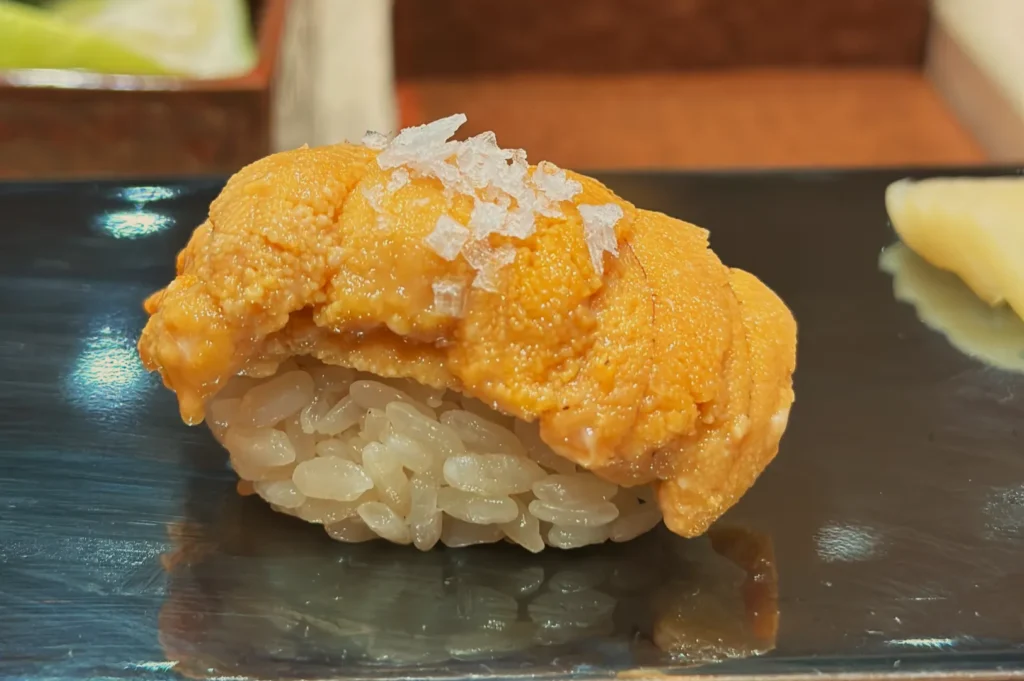 A piece of sushi featuring a mound of yellow sea urchin (uni) on rice, garnished with a sprinkle of salt, placed on a dark plate.