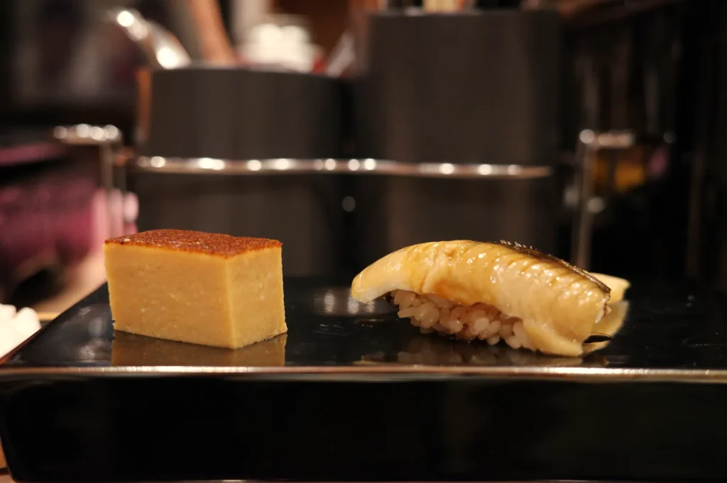 A plate featuring two pieces of sushi: one slice of tamago (Japanese sweet omelette) and one piece of sushi topped with mackerel.