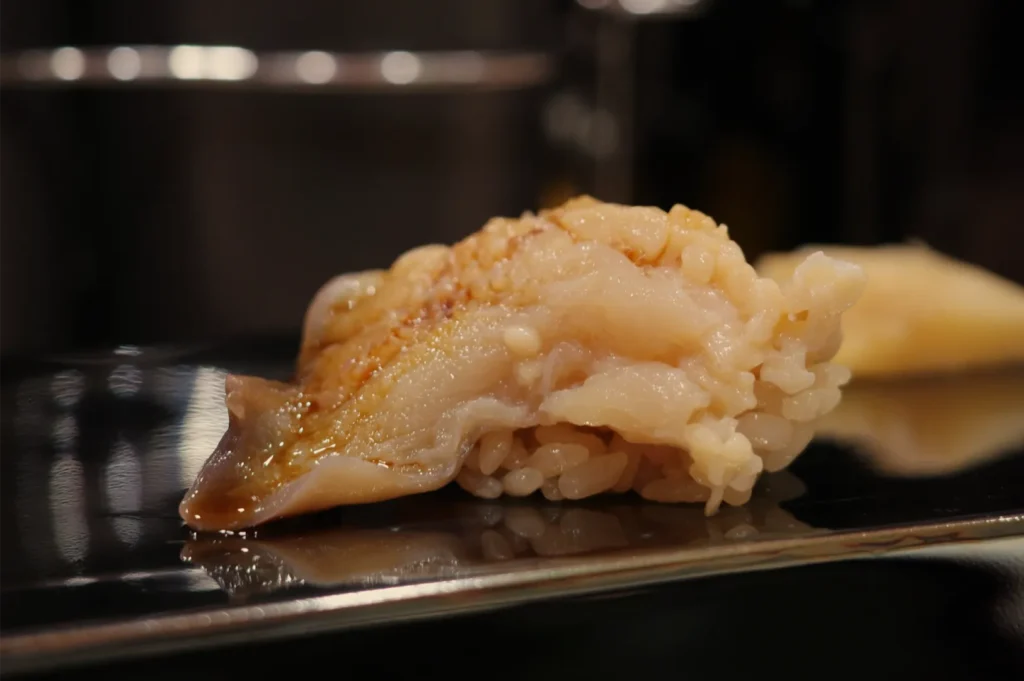 Close-up of a glistening piece of sushi, topped with tender grilled fish and rice, on a reflective black plate; warm, elegant dining mood.