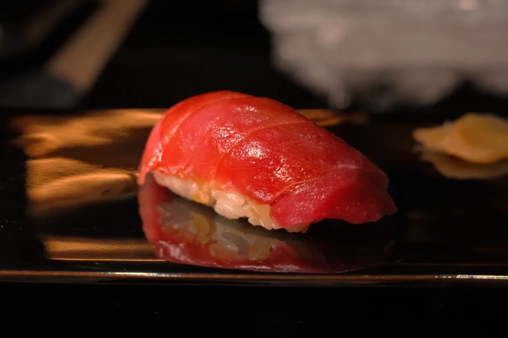 A piece of sushi with a vibrant red slice of fish on top, sitting on a black plate with a reflective surface.