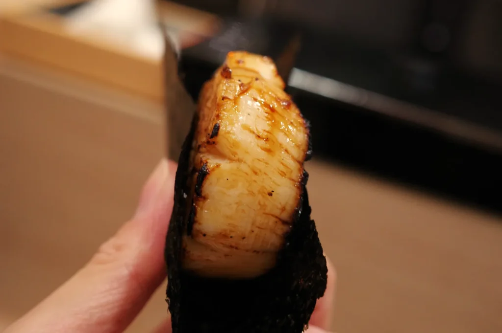 A close-up of a piece of sushi, featuring a shiny, grilled fish fillet wrapped in seaweed.
