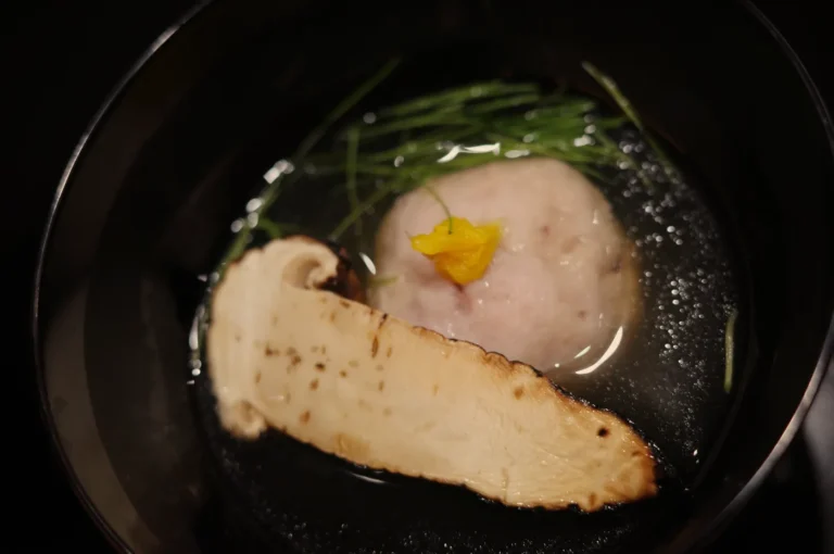 A close-up of a pale fish fillet and round fish cake simmering in a dark pan with green herbs and a small yellow garnish, evoking a quiet, rustic meal.