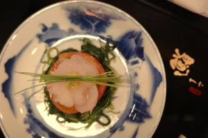A delicate seafood appetizer with pale shrimp on a red tomato slice, topped with chives and greens, served on a blue-and-white patterned plate.