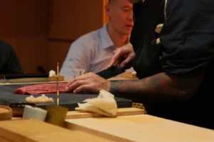 A tattooed Chef Masa in black cuts raw fish at a wooden sushi counter, with a customer watching in the warm, intimate restaurant setting.