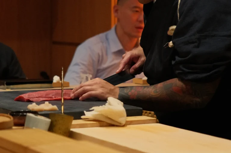 A tattooed Chef Masa in black cuts raw fish at a wooden sushi counter, with a customer watching in the warm, intimate restaurant setting.