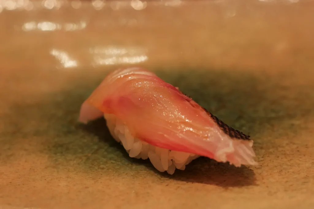 Close-up of translucent pink tuna nigiri sushi with glossy fish over white rice on a tan plate, warmly lit in a calm, appetizing setting.