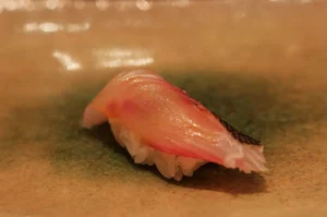 Close-up of translucent pink tuna nigiri sushi with glossy fish over white rice on a tan plate, warmly lit in a calm, appetizing setting.