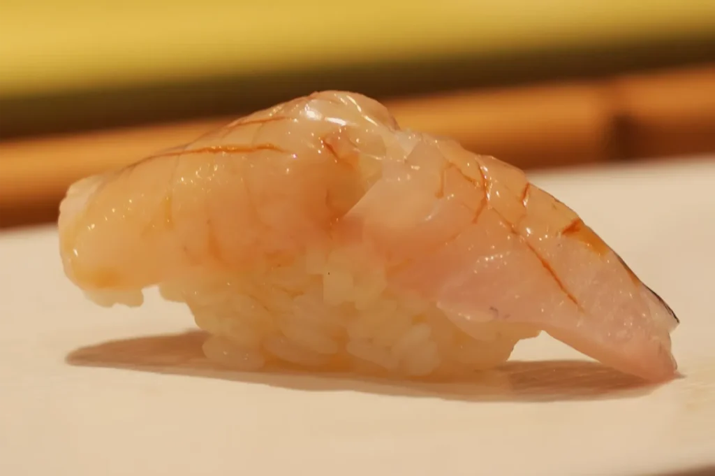 A translucent piece of raw shrimp rests on a pale plate, its glossy orange-pink flesh highlighted against a softly blurred warm background.