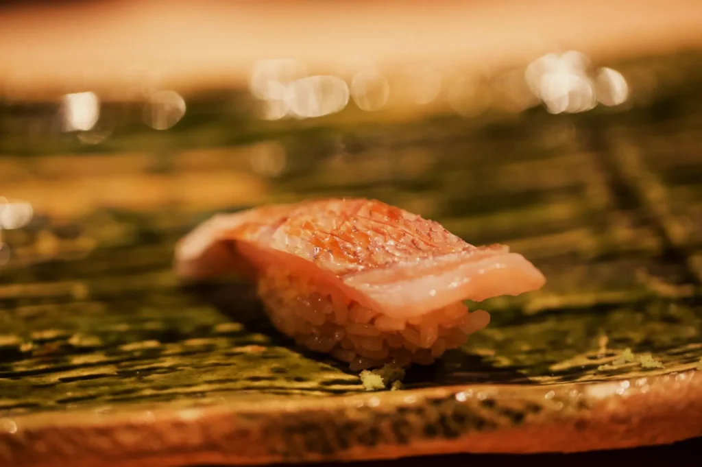 A slice of salmon nigiri rests on a dark green leaf in warm, golden light, with blurred lights behind it creating an intimate, appetizing mood.
