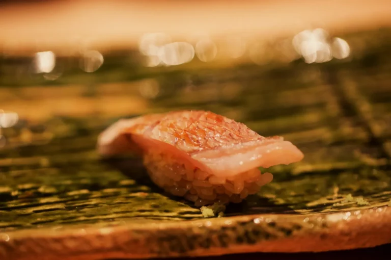 A slice of salmon nigiri rests on a dark green leaf in warm, golden light, with blurred lights behind it creating an intimate, appetizing mood.