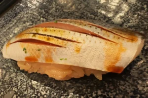 Close-up of seared salmon nigiri on a dark speckled plate, with browned scoring, orange edges, and rice beneath; warm and appetizing.