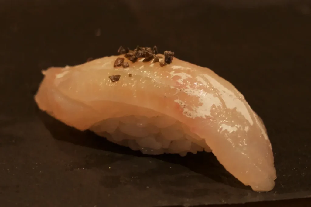 A glossy piece of salmon nigiri sushi topped with dark seasoning rests on a black plate, its warm lighting giving the food an appetizing, elegant look.